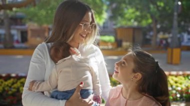 Anne bebeğini kucağında tutuyor ve güneşli şehir parkında genç kızıyla konuşuyor. Çiçekler etrafını sarmış, aile sevgisini ve açık hava ilişkisini tasvir ediyor..