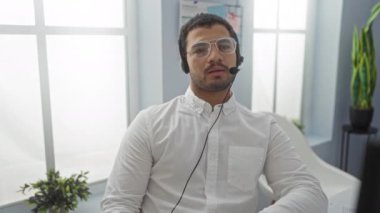 Kulaklık takan ve gözlüklü bir adam, modern bir ofiste bitki ve pencereli, bir sohbete katılmış, çağdaş bir iş yerinde uzaktan çalışan bir profesyoneli canlandırıyor..