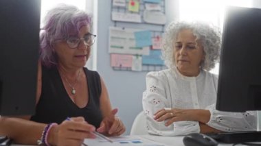 İki iş kadını bir ofis masasında bilgisayar ve evrak işleriyle uğraşıyorlar, profesyonel bir çalışma alanında yaptıkları toplantıda ortaklıklarını ve takım çalışmalarını vurguluyorlar.