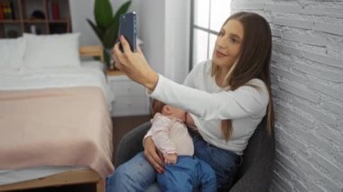 Kadın çocuğunu emzirirken rahat bir yatak odasında selfie çekiyor, anne-çocuk bağını kapalı bir yatakta yakalıyor, akıllı telefon ise modern aile anlarını simgeliyor..