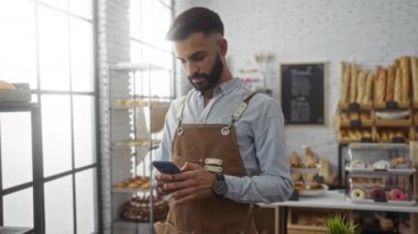Önlüklü genç sakallı adam raflarda çeşitli ekmek ve hamur işleri sergilenen bir fırında akıllı telefon kullanıyor..