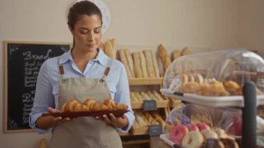 Pastanede elinde kruvasanlarla çalışan bir kadın. Önlük takıyor ve arka planda ekmek rafları ve hamur işleri var.