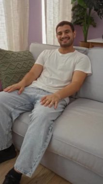 Genç İspanyol bir adam rahat bir oturma odasında kanepede oturuyor, gülümsüyor ve günlük giysiler giyiyor..