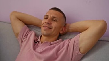 Oturma odasında pembe gömlekli ve gülümseyen bir ifadeyle rahatlayan genç adam, mor arka planlı bir ev ortamında yakalandı.