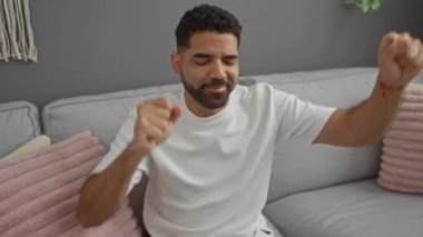 Oturma odasında kanepede otururken dans eden ve kollarını kaldıran mutlu bir adam.