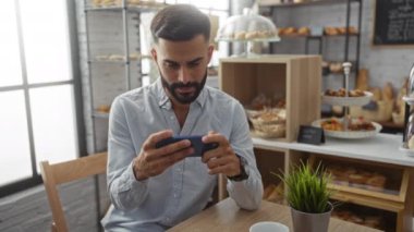 Sakallı genç adam fırında telefon kullanıyor. Arka planda hamur işleri ve bitkiler var. Kapalı alanda otururken odaklanmış görünüyor..