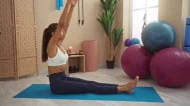 Spor salonunda yoga minderinin üzerinde uzanan genç bir kadın. Spor topları ve spor aletleriyle çevrili.