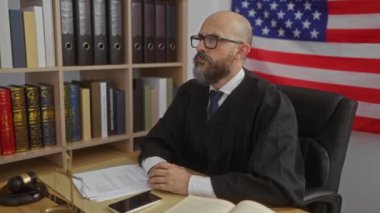 Bir ofiste oturan, arkasında Amerikan bayrağı olan hukuki bir cübbe giyen, etrafı hukuk kitaplarıyla çevrili, ve masanın üzerinde bir tokmak ve belgeler olan bir adam..