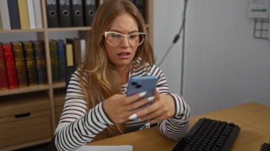 Ofiste akıllı telefon kullanan bir kadın. Gözlüklü ve sarı saçlı. Etrafı kitaplık ve masa eşyalarıyla çevrili.