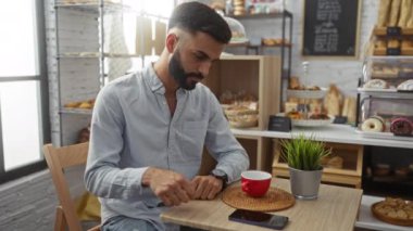 Fırın masasında bekleyen genç bir adam, kırmızı kahve fincanı ve masada bitkiyle telefonuna bakıyor, rahat bir kafe içinde fırın ürünleriyle çevrili, birini bekliyor..