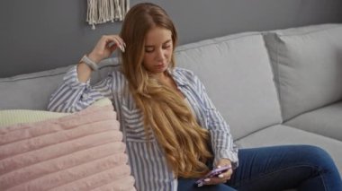 Genç bir kadın oturma odasında dinleniyor, akıllı telefonuyla bir koltukta oturuyor, çizgili gömlek ve kot pantolon giyiyor, sarı saçlı, içeride, evde oturuyor..