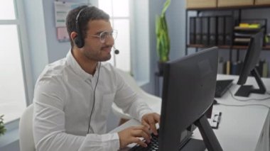 Ofiste çalışırken gülümseyen, kulaklık takan ve bilgisayarda yazan genç İspanyol bir adam.