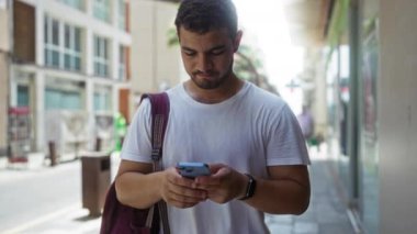 Kentsel sokak ortamında akıllı telefon kullanan, beyaz tişört giyen ve çanta taşıyan genç bir adam, ekrana odaklanmış, binalar ve diğer kentsel unsurlarla çevrili.