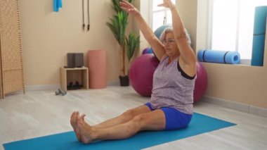 Spor salonunda yoga yapan yaşlı bir kadın paspas ve denge topları gibi egzersiz ekipmanlarıyla dingin bir ortamda zindelik ve sağlığını gösteriyor.