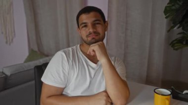 Beyaz tişörtlü, arka planda siyah perdeleri ve sarı fincanı olan, düşünceli bir ifadeyle bir oturma odasında oturan İspanyol genç adam.