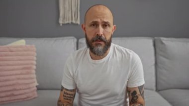 Dövmeli ve sakallı bir adam oturma odasında beyaz tişört giyerek oturuyor.