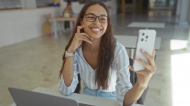 İspanyol kadın ofiste dizüstü bilgisayarla oturmuş gözlüklü ve çizgili tişörtlü bir şekilde selfie çekiyor.