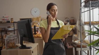 Bir pastanede elinde pasta rafları ve yanında kurabiye kavanozu olan bir tezgahla elinde bir panoyla telefonla konuşan bir kadın.