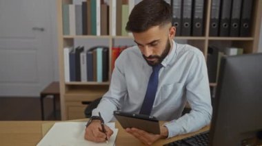 Ofiste çalışan yakışıklı İspanyol bir adam tableti inceliyor ve çalışma odasına kitaplıklarla notlar yazıyor.