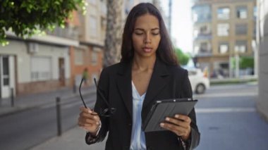 Şehir caddesinde gözlüklü bir kadın tablet okuyor. Arka planda binalar ve arabalar var. İş dünyası ve teknoloji sergileniyor.
