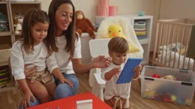 Kadın ve iki çocuğu rahat bir kreşte, oğlu tablet kullanıyor ve kızı kucağında oturuyor, etrafı oyuncaklarla çevrili, beşikte, aile sevgisini ve bağlantısını gösteriyor..