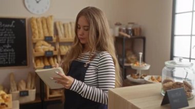 Fırın dükkanının içindeki not defterine yazan kadın etrafı fırınlanmış mallar, saatler ve karatahta menüleriyle çevrili.