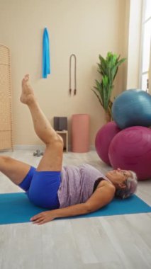 Spor salonundaki yoga minderinde bacak kaldırma egzersizi yapan arka planda egzersiz topları ve yeşillik olan bir kadın.