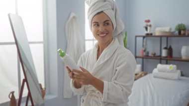 Parlak, dingin bir odada kafasında bir havluyla bir sağlık spasında losyon şişesini tutarken gülümseyen bir kadın..
