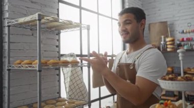 Fırında çalışan genç bir adam, arka planda tatlılar olan, büyük pencereli parlak bir odada raftaki taze pişmiş kurabiyeleri kontrol ediyor.