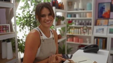 Bir dekor mağazasında elinde akıllı telefon tutarken kameraya gülümseyen bir kadın. Etrafı çeşitli ürünlerle çevrili iç mekan bitkileri ve raflarla çevrili.