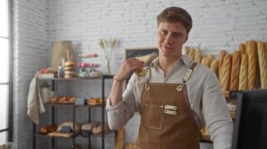 Önlüklü genç adam ekmek ve hamur işleriyle dolu bir fırında güvenle duruyor..