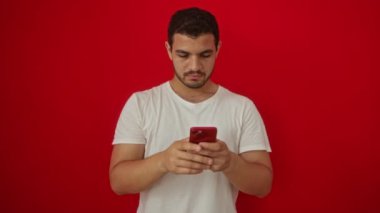 Genç İspanyol adam, izole edilmiş kırmızı arka plan üzerinde akıllı telefon kullanırken ciddi bir hareketle seni işaret ediyor.