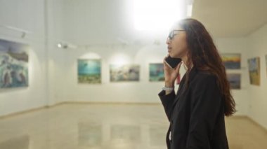 Tablolarla çevrili bir sanat galerisinde telefonla konuşan, geniş ve aydınlık bir ortamda meşgul ve odaklanmış bir konuşma yapan bir kadın..