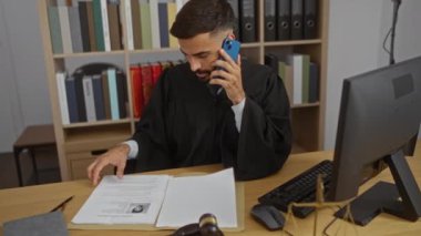 Yargıç cüppeli genç adam ofis masasında bilgisayar monitörüyle yasal belgeleri incelerken cep telefonuyla konuşuyor.