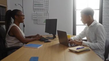 Ofiste gülümseyen kadın ve adam, duvarında dokümanlar ve çizelgelerle dizüstü bilgisayarlar ve bilgisayarlarla çalışırken yumruk tokuyor.