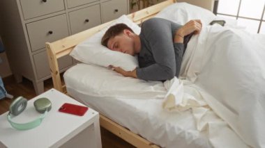 Yatakta akıllı telefonlu bir adam rahat yatak odasındaki yan masada uyur. Hafif duvarları, ahşap mobilyaları ve nötr dekoru vardır. Huzurlu bir ortamda huzur içinde dinlenir.