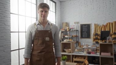 Önlüklü genç adam pastanede duruyor. Arka planda ekmek ve pastalar var.