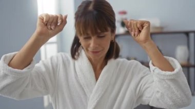 Kısa esmer, İspanyol bir kadın sıcacık bir sağlık merkezinde spa gününün tadını çıkarıyor, beyaz bir bornoz giyiyor ve neşe saçıyor..