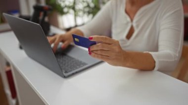 Sarışın, evde oturan yaşlı bir kadın elinde mavi bir kredi kartı olan dizüstü bilgisayarını kullanarak online alışverişi ön plana çıkaran ev dekorasyonu malzemeleriyle çevrili bir odada oturuyor.