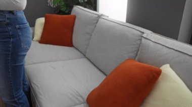 Gri bir kanepede, rahat bir oturma odasında turuncu yastıklarla dinlenen bir kadın..