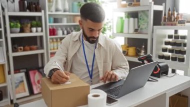 Genç adam bir ev dekorasyonu mağazasında çalışıyor, bir paketin üzerine dizüstü bilgisayar ve barkod tarayıcısı yazıyor, çeşitli dekoratif malzemelerle çevrili..