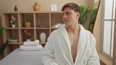 Beyaz cüppeli genç adam, bitki ve havlularla dolu bir spa odasında dinleniyor, huzurlu bir sağlık deneyimi yaşıyor.