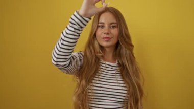 Çizgili tişört giyen genç sarışın kadın, sarı arka planda izole edilmiş parmaklarının arasından bakarken mutlu bir gülümsemeyle el üstünde işaret yapıyor.