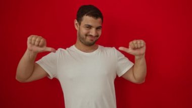 Beyaz tişörtlü, kendine güvenen genç İspanyol adam kendini göstererek izole edilmiş kırmızı arka planda mutlu ve pozitif görünüyor.