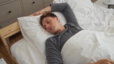 Genç adam, beyaz yorganın altında, arka planda tahta yatak başlığı ve dolabı olan uzun kollu gri bir gömlek giyerek huzur içinde uyuyor.