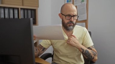 Adamın biri çalışma ortamında kendini belgelerle körüklüyor. Masasında otururken stresli ve rahatsız görünüyor.