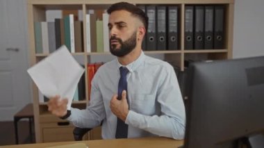 Ofisteki genç adam, arka plandaki raflarda kitap ve klasörlerle masasında otururken kendini kağıtlarla yelpazeliyordu.