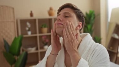 Sağlık merkezindeki genç adam, bitki ve raflarla çevrili beyaz bir bornoz giyerek cildini inceliyor.