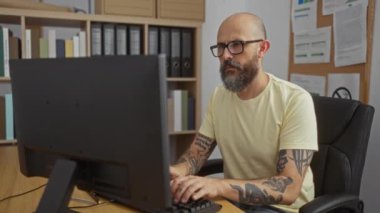 Ofiste bilgisayar başında çalışan, sarı gömlekli, sakallı ve dövmeli İspanyol bir adam kolunu acıdan tutuyor.