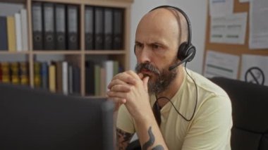 Sakallı ve dövmeli bir adam ofiste çalışıyor. Kitaplar ve klasörler arka planda.